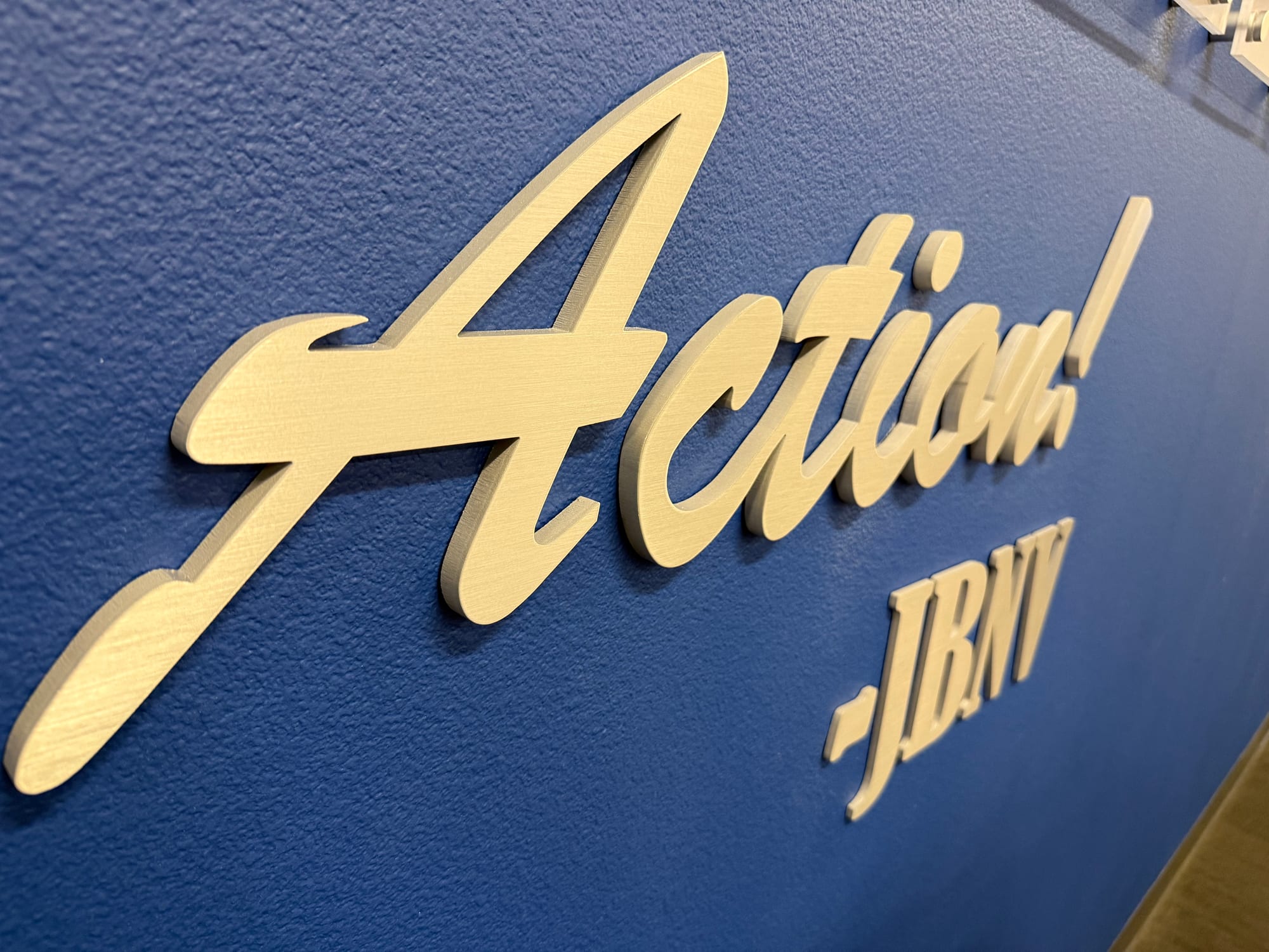 custom metal sign gold brushed letters las vegas Custom metal sign: “Action! JBNV” in gold brushed metal script letters on a blue wall